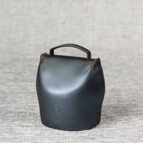 Cloche de vache métallique gris foncé et patinée, posée droite sur un tissu texturé gris clair. Elle porte le logo d'une tête d'animal, « PREMANA » et « 10 » gravés.