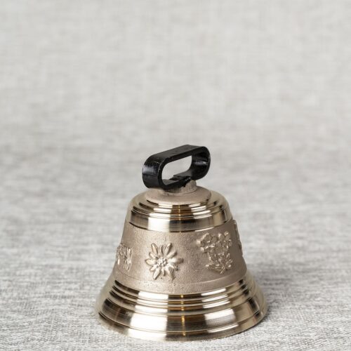 Cloche décorative en bronze, ornée de motifs floraux dont des edelweiss, posée verticalement sur un tissu gris clair texturé.