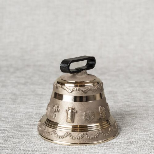 Cloche de vache miniature en bronze, ornée de motifs ruraux (pot à lait, fleurs) et d'une poignée noire, reposant sur une surface textile gris clair.