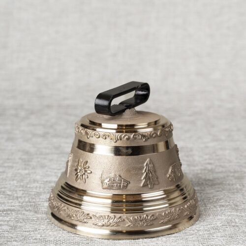 Cloche décorative en bronze ornée de motifs alpins (édelweiss, chalets, sapins) et d'une anse noire, posée sur une surface textile gris clair.