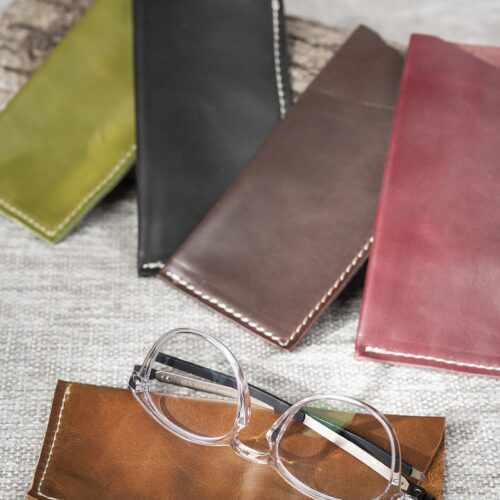 Des lunettes à monture transparente (CE 2019 XENCE) posées sur un étui en cuir marron. Derrière, d'autres étuis colorés (vert, noir, marron, rouge) sont alignés sur un tissu gris et un morceau de bois.